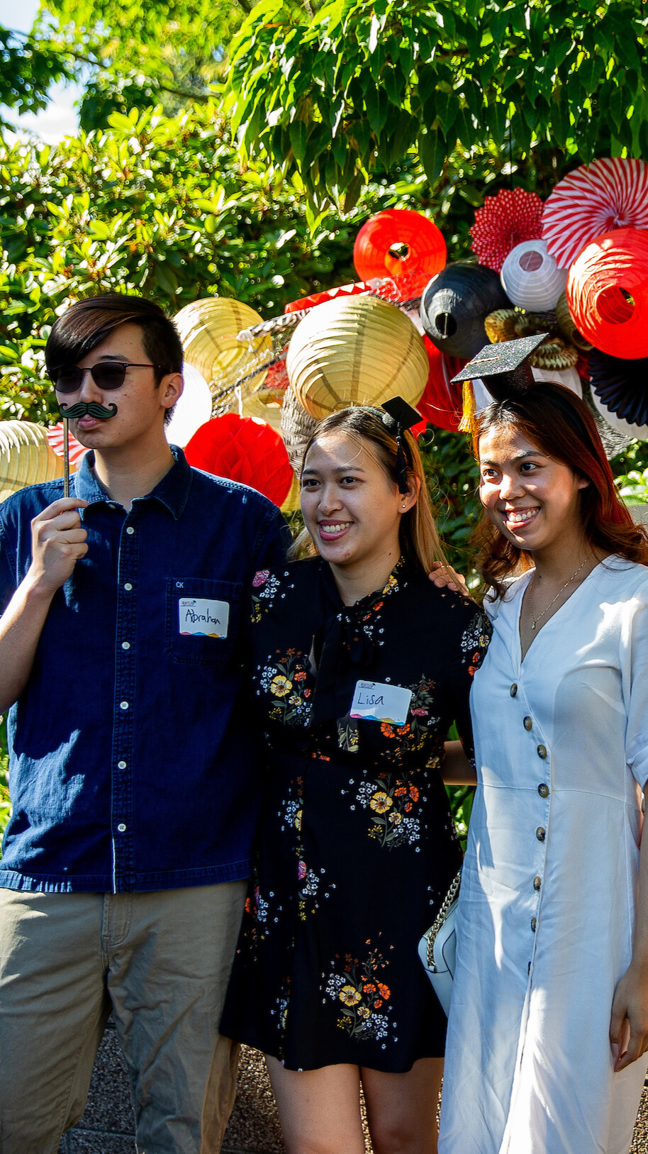 Three people poses with silly props in front of a balloon arch.