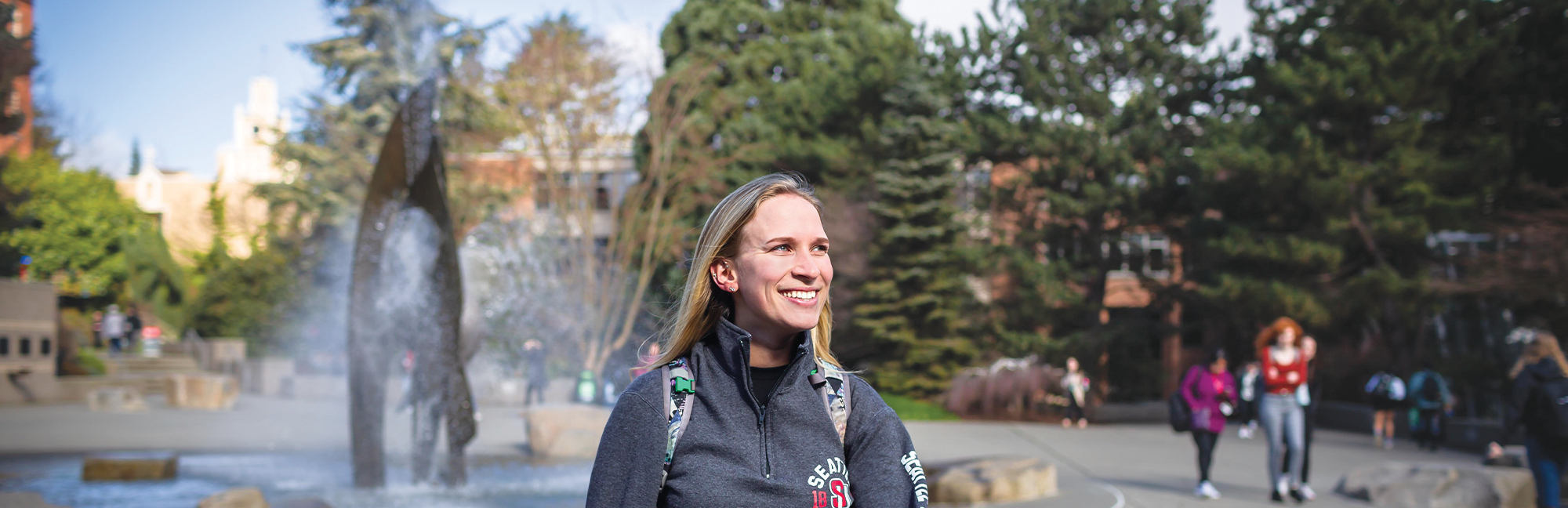 A student wearing Seattle University gear walks through the campus quad with the fountain in the background.